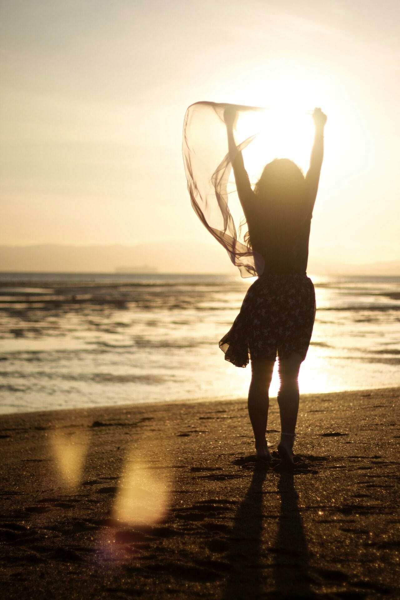 A woman with her arms raised standing on a beach by the shore